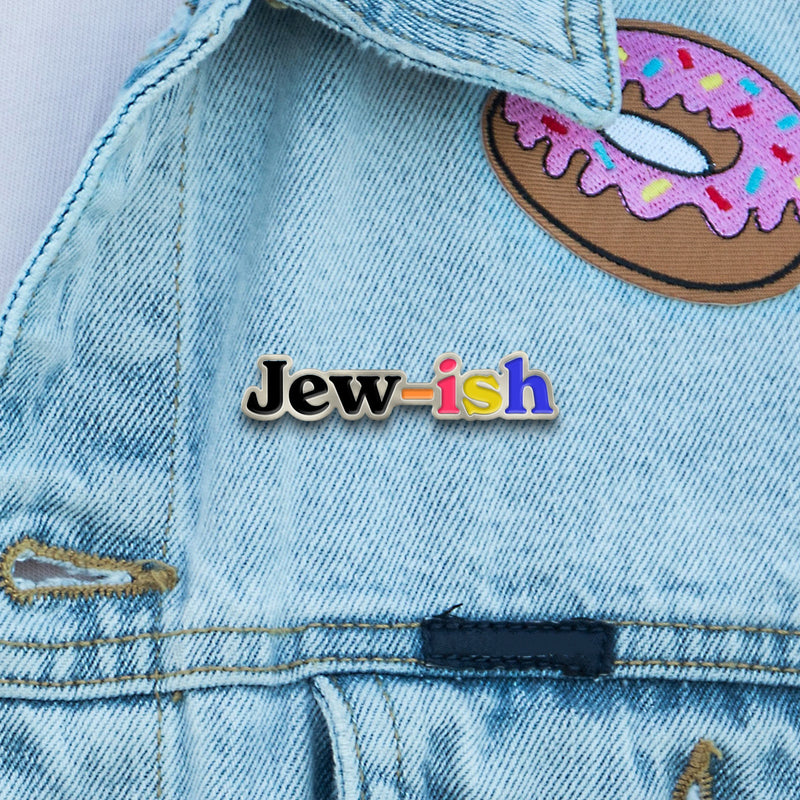 multicolored silver enamel pin that reads "jew-ish", pinned into a denim jacket where a donut-designed patch is also shown
